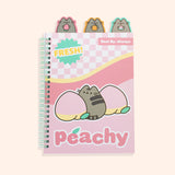 A light pink spiral bound notebook features Pusheen between two pink and yellow peaches. Extending off the top of the colorful peach notebook are three tabs featuring a grey Pusheen the cat holding a strawberry, orange, and watermelon slice. The front of ntoebook features phrases including “fresh!” “best by: always,” and “peachy.”
