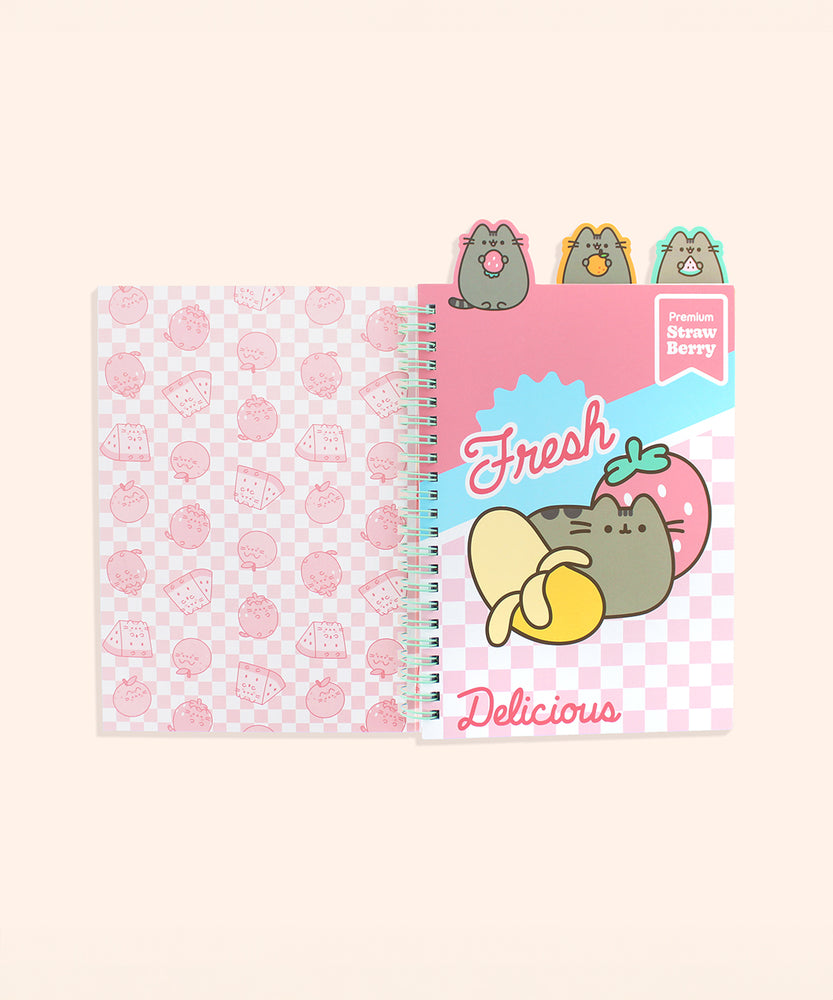 The cover of the notebook opened to the first page. The interior cover features a pink all over pattern of Pusheen as various Fruit characters in front of a pink checkered background. The first page has a pink patterned background with a graphic of Pusheen with a partially peeled banana and a pink strawberry surrounded by the phrases “fresh,” “premium strawberry,” and “delicious.: The pattern page is attached to the tab of Pusheen holding a pink strawberry.