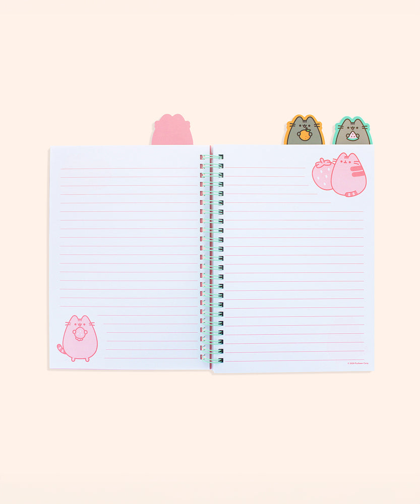 The open notebook shows a pink lined page with a pink Pusheen and strawberry in the top left corner. Above the notebook are two tabs featuring Pusheen holding an orange and watermelon slice. The left page has a graphic of Pusheen standing while holding a strawberry upside down in the bottom left corner.