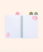 The open notebook shows a pink lined page with a pink Pusheen and strawberry in the top left corner. Above the notebook are two tabs featuring Pusheen holding an orange and watermelon slice. The left page has a graphic of Pusheen standing while holding a strawberry upside down in the bottom left corner.
