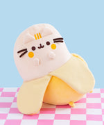 Front view of the Pusheen Fruits Scented Banana Squisheen Plush. The yellow plush sits upright on a pink and white checkered surface. The plush resembles a partially peeled banana.
