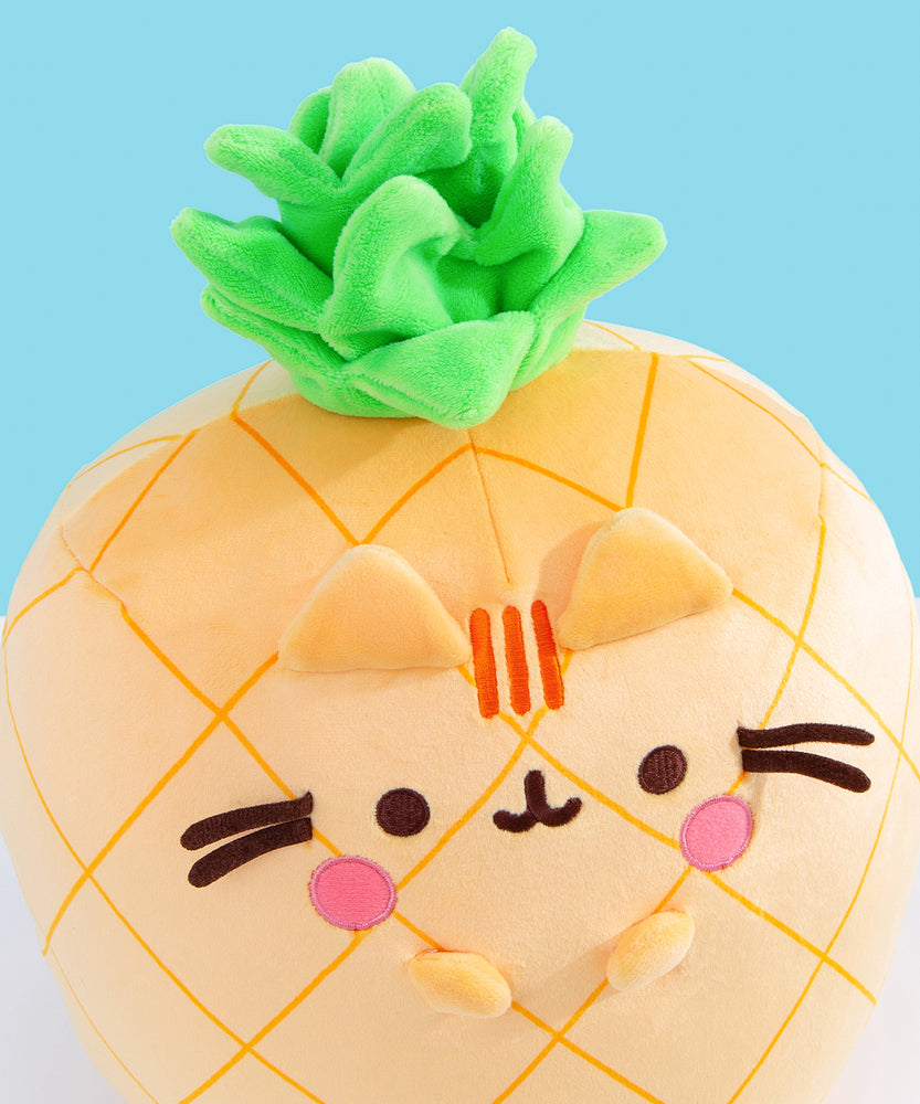 Aerial view of the Pineapple plush. The vibrant green crown has different layers of leaves to mimic a real pineapple stem. Pusheen’s face includes her classic three head stripes, eyes, mouth and whiskers plus light pink blush spots near her mouth. 