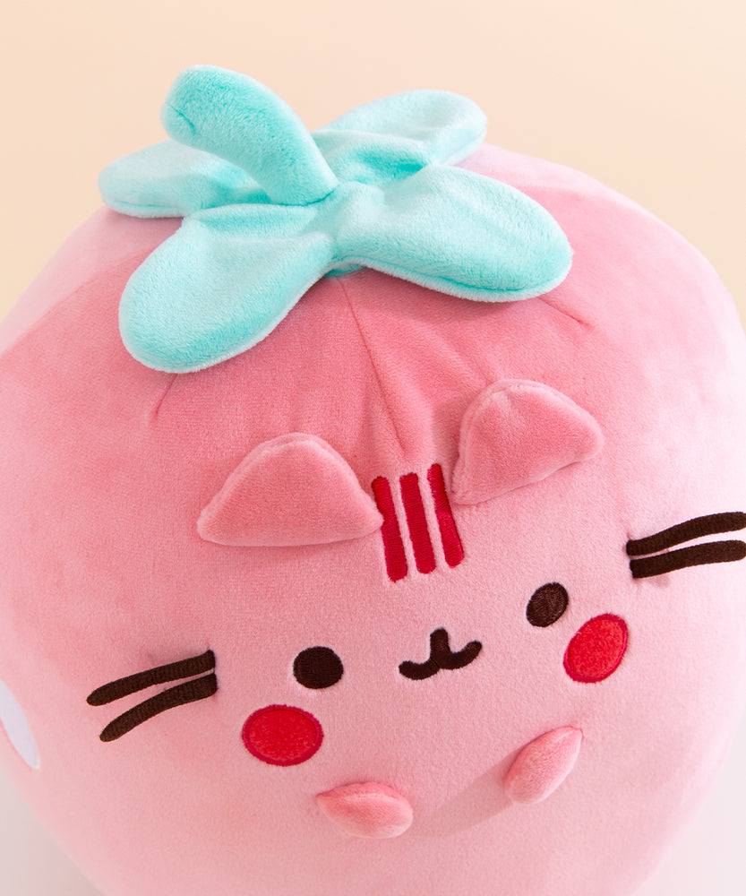 Aerial view of the Strawberry plush. The light green stem has four leaves to mimic a real strawberry. Pusheen’s face includes her classic three head stripes, eyes, mouth and whiskers plus light pink blush spots near her mouth. 
