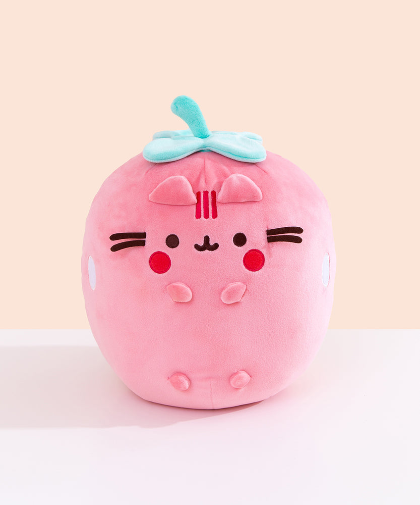 Front view of the Pusheen Fruits Scented Strawberry Squisheen Plush. Pusheen the Cat is a light pink strawberry with a mint green stem and leaves that’s sitting atop a white tabletop.  