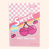 Front view of the notebook in the Pusheen Fruits Stationery Set. The pink notebook has a graphic of Pusheen as two pink cherries on stems. Phrases included on the front of the notebook include “sweet,” “fresh!,” “open,” and “100% real cute.” The background of the notebook has a pink checkered print with pink and yellow stripes.