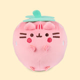 Front view of the Pusheen Fruits Strawberry Squisheen Plush. The light pink plush stands 4” tall and has Pusheen the Cat’s facial features embroidered on the front of the rounded form in dark brown and dark pink. Pusheen is blushing and smiling. Coming off the top of the plush is a light green stem and leaf felt piece. 