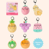 Front view of Pusheen Fruits Surprise Plush assortment. Pusheen the Cat has taken the form of seven miniature fruit-inspired plushes. At the top left is the packaging box for the Pusheen Fruits surprise plush. From left to right and top to bottom, the surprise plush are a strawberry, half-peeled banana, pineapple, grape cluster, watermelon slice, cut kiwi, and an orange. The bottom right corner has a burst with the words “1 Mystery Design” indicating eight possible miniature plush keychains are available. 