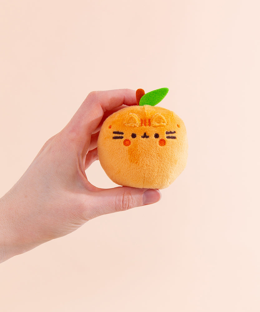 Model holds orange fruit with Pusheen’s facial features. The orange plush body has a brown stem and green leaf coming out the top of the plush keychain. 