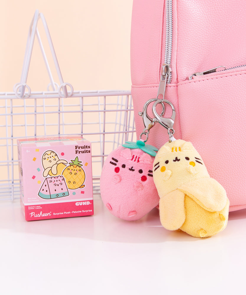 Close-up shot of Pusheen Fruits characters: strawberry and banana. The plush keychains are attached to a pink bag. Next to the yellow and pink plushes is the fruits surprise plush packaging box. 