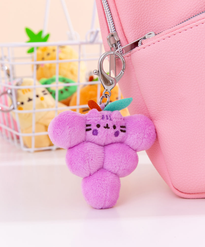 Close-up shot of Pusheen Fruits character: grapes. The purple cluster of grapes plush keychain is attached to a pink bag with the various other fruits character keychains shown in the background.  
