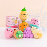Front view of Pusheen Fruits Surprise Plush assortment. Plush keychains sit on multi-level white pedestal in front of a light-yellow background. Keychains feature various fruits including a pineapple, strawberry, orange, kiwi, grapes cluster, watermelon slice, and peeled banana. Accompanying the seven fruits keychains is the mystery box packaging for the Fruits Surprise Plush. 
