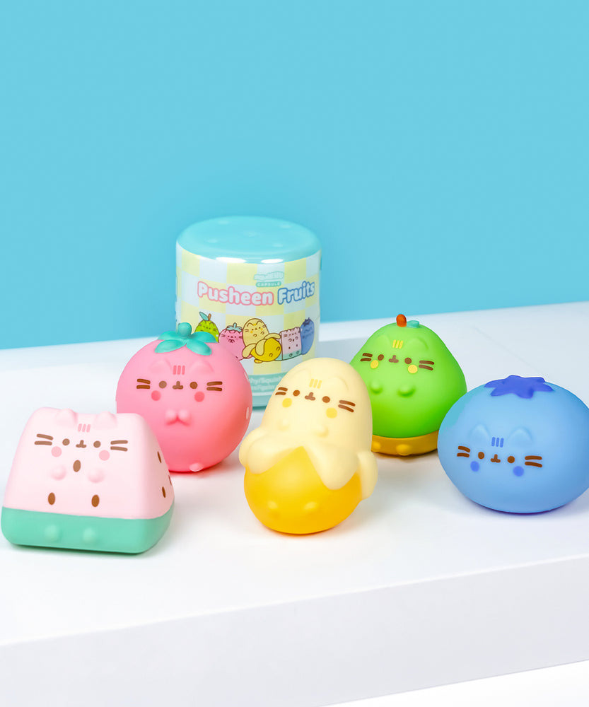 Front view of all Pusheen Fruits Surprise Squishy including the blind box capsule. The Squishys include a pink and mint watermelon slice, pink strawberry, yellow partially-peeled banana, green and brown pear, and blue blueberry.