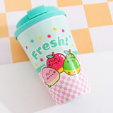 The Pusheen Travel Mug lies on a white and yellow checked surface. The lid comes with a stopper molded in the lid that allows the hold to be opened and closed. 