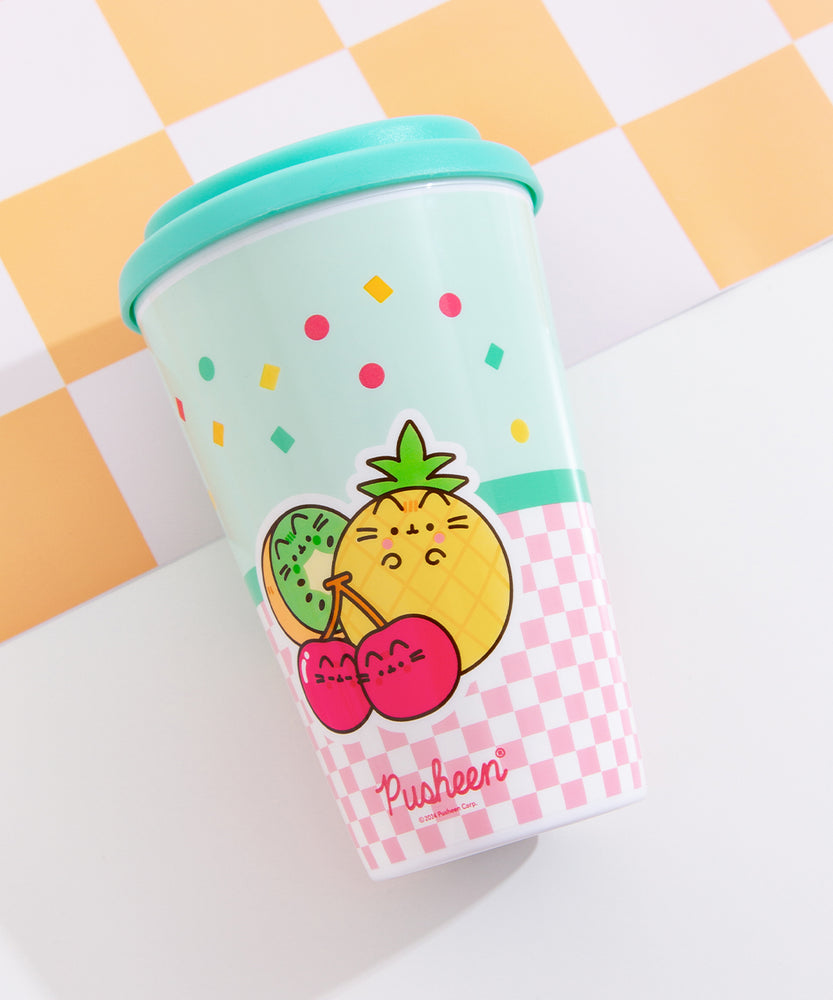 The travel mug lies on its front to show the printed back graphic. The graphic includes three Pusheen Fruits characters including a green and brown kiwi slice, a checkered yellow pineapple, and a set of red cherries on stems.  