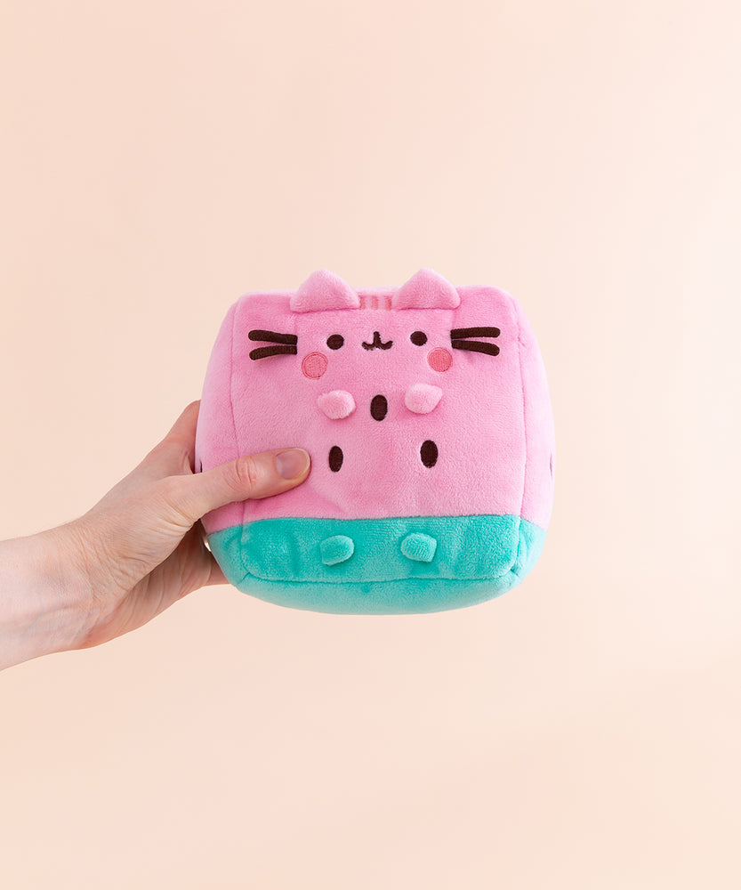Model holding side of the Pusheen Fruits Watermelon Plush. Pusheen the Cat takes the form of a juicy pink and mint green watermelon slice. The triangle shaped plush body has embroidered brown cylinder shapes to represent watermelon seeds. On the front of the plush are 3D versions of Pusheen’s ears and four paws.  