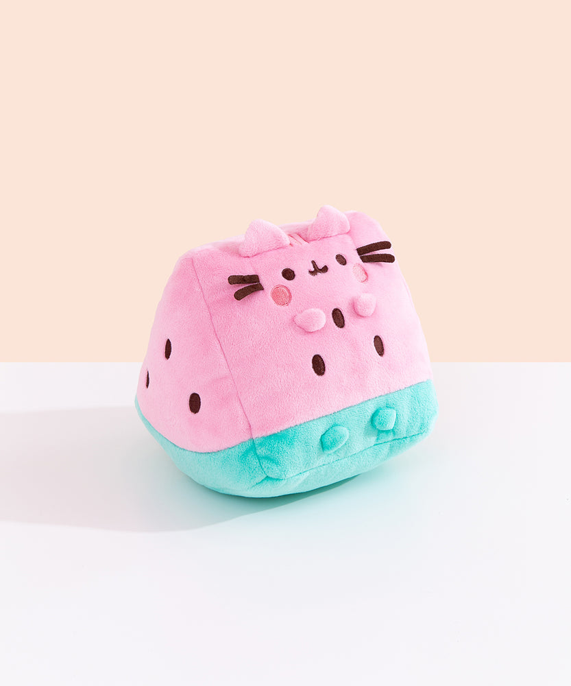 Side quarter view of the plush. The plush sits upright while Pusheen’s four paws extend off the front of the plush and do not touch the tabletop surface the plush is sitting on. The brown embroidered spots on the sides and front of the plush mimic watermelon seeds. 