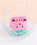 Pusheen Fruits Watermelon plush sits inside a white metal carrying basket. The pink and green plush has brown and darker pink embroidery details.  