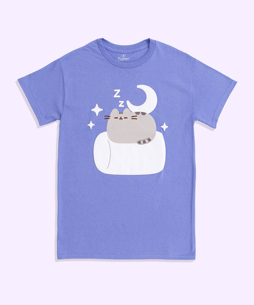 A periwinkle graphic t-shirt lies against a light purple background. The unisex tee shirt has a screen-printed graphic in the center of the chest showing Pusheen the Cat resting on a large, white marshmallow surrounded by a white crescent moon, stars, and zzz’s. 