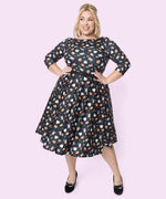 Model wears Pusheen Halloween Dress. The black retrofit dress has three-quarter length sleeves and a flowy skirt that falls below the knee. The dress has an allover print of a white Boosheen and a grey Witch Pusheen wearing a purple hat and riding a broomstick. 