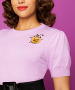 Close-up of Pusheen and Jack-O-Lantern embroidery on wearer’s left chest. A greyPusheen sits in an orange and black Jack-O-Lantern with a top of the pumpkin and stem sitting on her head as a hat. The cat is surrounded by five black stars. 