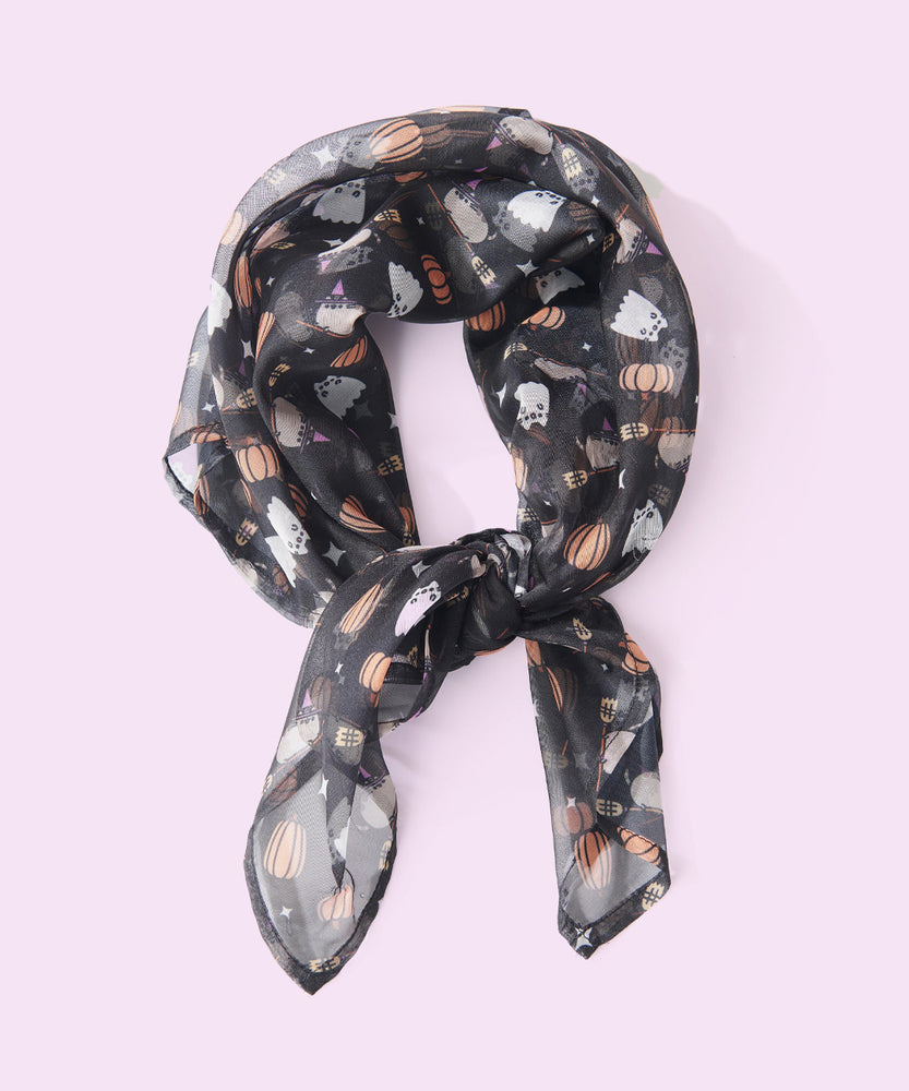 Pusheen Halloween Scarf lies on a light purple background tied in a knot to show the circumference of the scarf when tied. The chiffon-style scarf has an allover print in grey, white, purple, brown, yellow, orange, and pink. 
