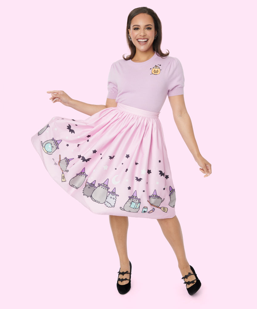 Front view of pink retro-inspired skirt featuring spooky bats and stars a repeating pattern graphic of Witch Pusheen, Witch Pip, and Witch Stormy at the bottom hem. The model’s hand holds skirt fabric away from the body to show off the skirt pattern. The Witch cats are wearing purple hats and Witch Pusheen rides a broomstick. 