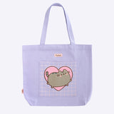 A tote bag is posed in front of a purple background. The purple tote has an appliquè of Pusheen the Cat blushing and jumping on the front pocket. The handles of the bag are purple, and the top has the Pusheen the Cat logo in pink and brown. 