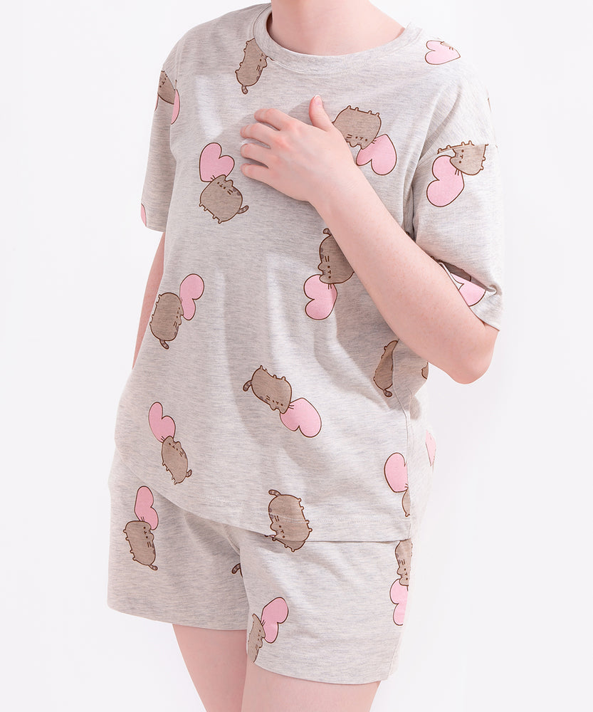 Model wears Pusheen Hearts Pajama Set. The two-piece loungewear set includes oversized grey top and elastic band shorts. Covering the top and shorts is a randomized pattern print of Pusheen and a light pink heart. 