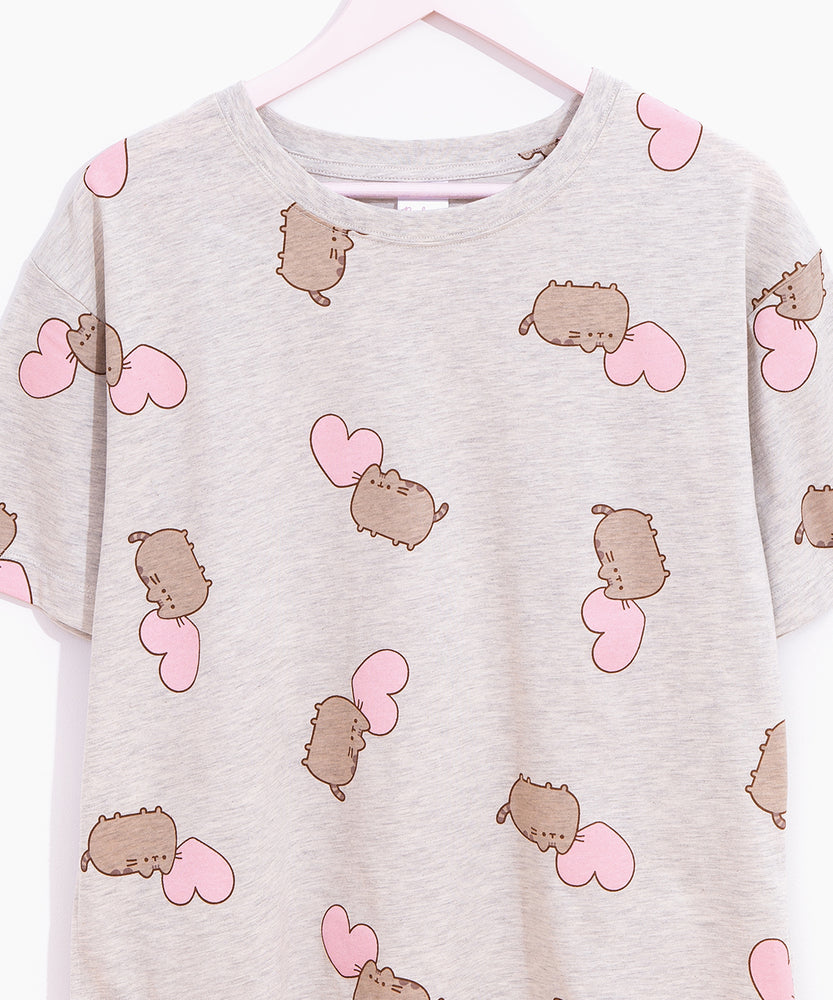 A close-up view of the pajama tee hung on a hanger in front of a white background. The gray pajama tee features a random pattern of Pusheen and pink hearts. 