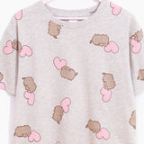 A close-up view of the pajama tee hung on a hanger in front of a white background. The gray pajama tee features a random pattern of Pusheen and pink hearts. 