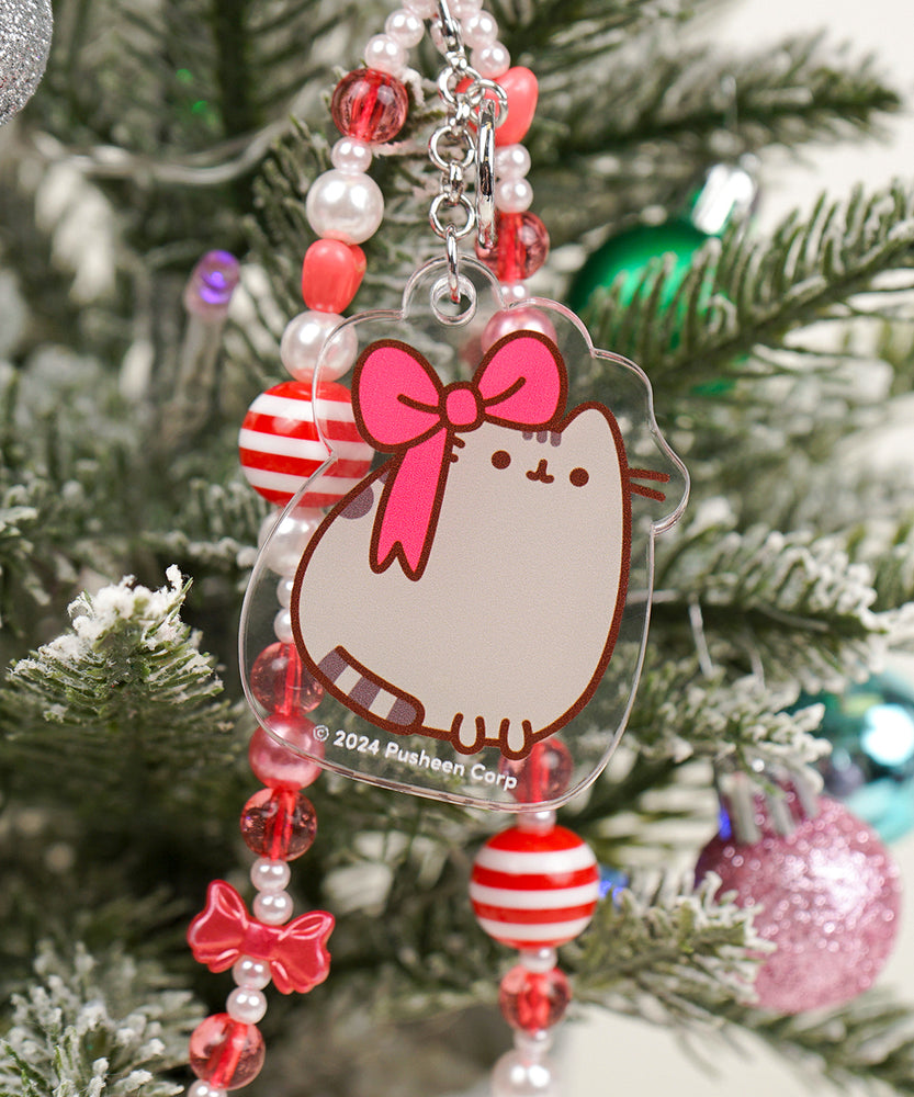 Close-up of Pusheen Holiday charm. The acrylic charm has a brown and grey tabby cat wearing a big red bow on one ear. 