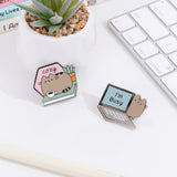 A close-up view of the two Pusheen pins lying on a white desk surrounded by a cactus plant and white keyboard. The book pin features a grey Pusheen the Cat with a green plant and book and a pink sign. The laptop pin features a grey laptop with light blue screen.  