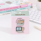 Pusheen Indoorsy Pin Set lies in front of a white desk with a keyboard, pen, and three Pusheen the Cat books. The Two Pusheen pins are attached to a backer card with the product name in dark brown color. The top pin shows Pusheen curled up with a mug on a green book. Behind the cat is a cactus plant and a sign that says “cozy.”  The bottom pin shows Pusheen peeking out from behind a laptop where the screen says, “I’m busy.” 