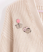 A close-up view of the two Pusheen pins attached to a cream knit cardigan. Pusheen is floating with the help of a pink balloon and the grey cat is also shown blushing while in a rounded pose. 