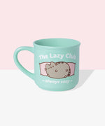 Front view of the Pusheen Lazy Club Mug. The rounded mint green mug is perfect for all hot beverages. The exterior has a mint green handle with matching interior walls. The outside of the mug features Pusheen the Cat lounging on two pink pillows with the saying “The Lazy Club” above the cat and “always cozy” below the cat graphic. 