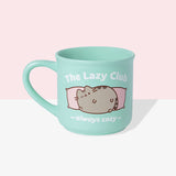 Front view of the Pusheen Lazy Club Mug. The rounded mint green mug is perfect for all hot beverages. The exterior has a mint green handle with matching interior walls. The outside of the mug features Pusheen the Cat lounging on two pink pillows with the saying “The Lazy Club” above the cat and “always cozy” below the cat graphic. 