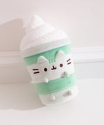 Close-up of Matcha Lattee Pusheen. The medium-green body is accompanied by light green horizontal lines to imitate a classic drink container. Pusheen's head stripes, eyes, smiling, mouth and whiskers are embroidered on the front of the plush while her light green ears and paws extend off the plush body.