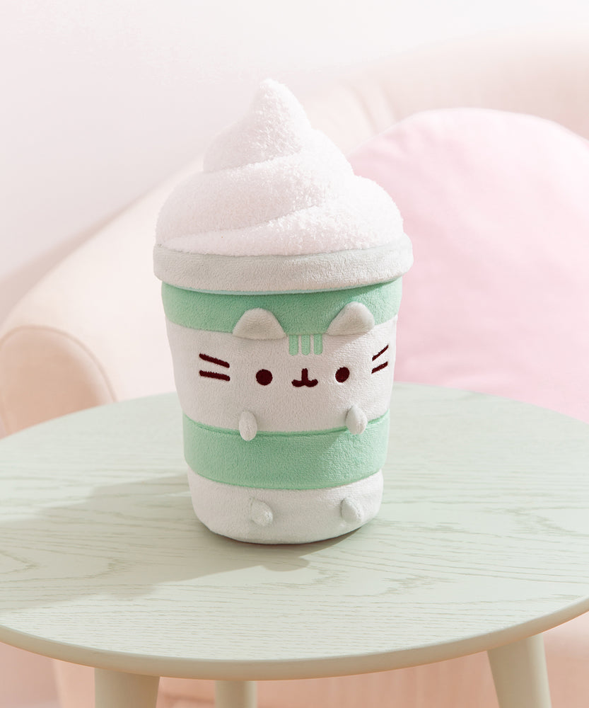 Matcha Latte Plush sits on a light green table top. Pusheen the Cat shows her love for the healthy green drink by becoming one as a plush.