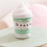 Matcha Latte Plush sits on a light green table top. Pusheen the Cat shows her love for the healthy green drink by becoming one as a plush.