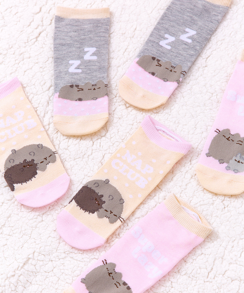 Pusheen Nap Club Socks lie on a light pink surface. The sock set features the medium grey Pusheen the Cat with her siblings: a small gray cat named Stormy and a dark brown cat named Pip. 