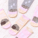 Close-up view of the graphics on the sock sets. The lefthand sock set features Pusheen and Pip napping on a yellow and pink sock with the phrase “Nap Club” in white text above the cats. The righhand sock set features Pusheen napping on a pink pillow on a grey and yellow sock with white “zz’s” above Pusheen.  