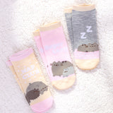 Close-up view of all three sock sets from the Pusheen Nap Club 3-Pack Ankle Socks that are laid flat in pairs on a white fluffy surface. From left to right are the yellow and pink “nap club” ankle socks, the “super lazy” pink and light-yellow sock set, and the light grey, pink, and yellow “ZZ's: sock set.  