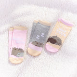 From view of the Pusheen Nap Club 3-Pack Ankle Socks. All three sock sets lay on a white fluffy surface with a ray of sunshine shining on the socks to show off the neutral colors. Each sock features a different Pusheen graphic.  