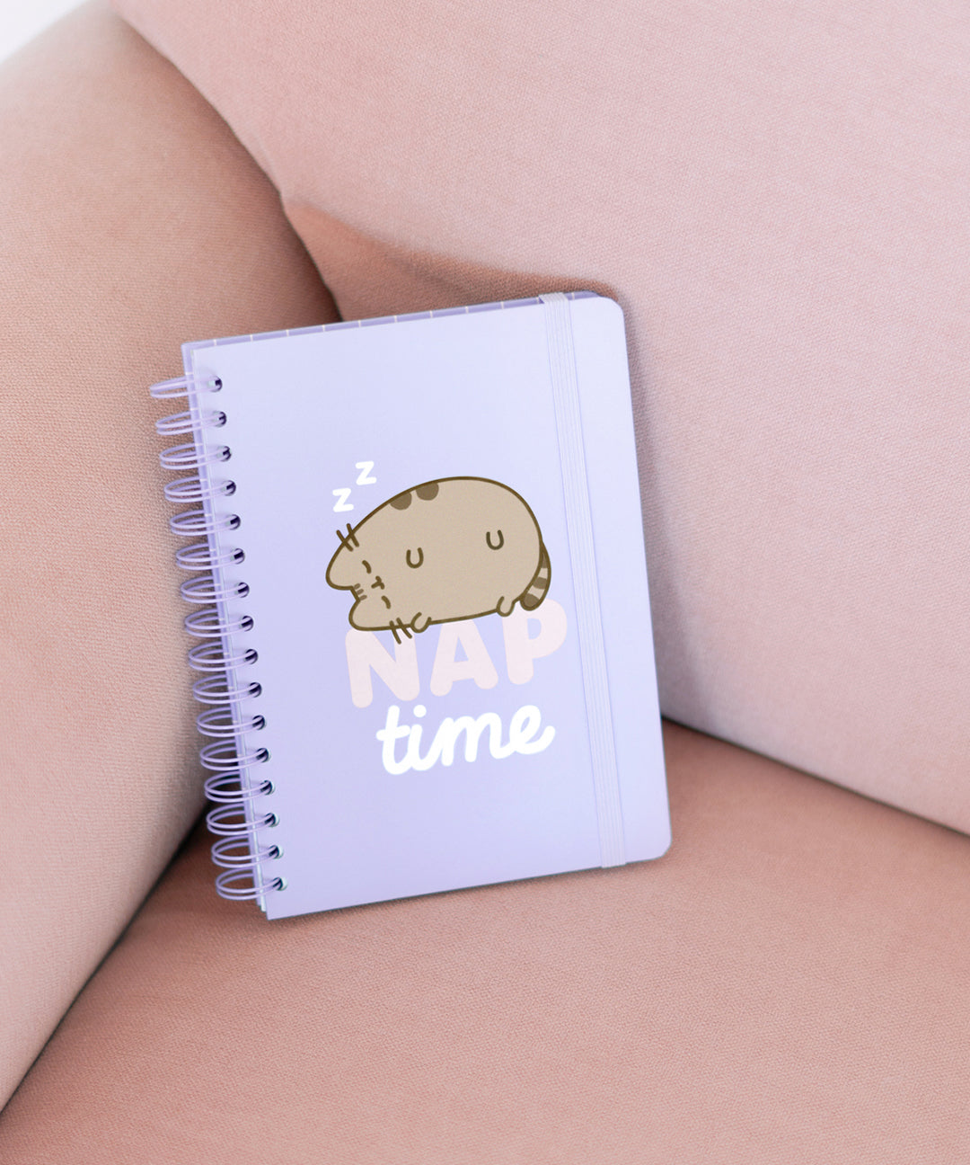 Front cover of the Pusheen Naptime Notebook. On the center of the light purple spiral notebook is a graphic of Pusheen the Cat sleeping on top of the phrase "nap time."