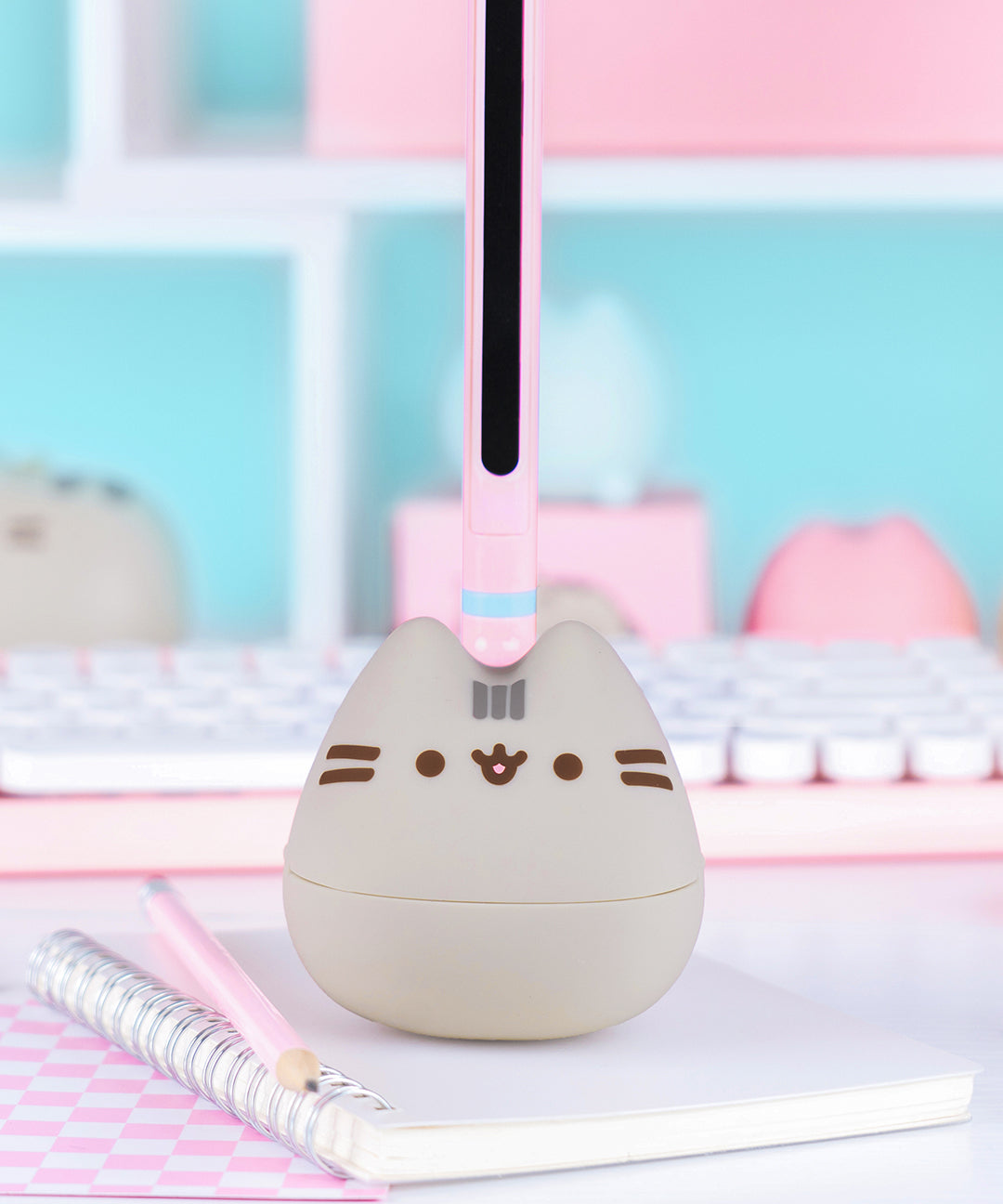 Otamatone sits on a desk. The pink and mint green stem has a black inner portion that allows the player to change the tone of the sounds coming out of the Pusheen Otamatone.
