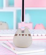 Otamatone sits on a desk. The pink and mint green stem has a black inner portion that allows the player to change the tone of the sounds coming out of the Pusheen Otamatone.