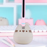 Otamatone sits on a desk. The pink and mint green stem has a black inner portion that allows the player to change the tone of the sounds coming out of the Pusheen Otamatone.