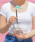 Model holds the Pusheen Otamatone. The synthesizer toy has a long stem with a musical note shaped top. The bottom portion is a round gray form that resembles Pusheen the Cat with her rounded triangular ears.