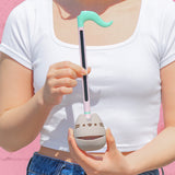 Model holds the Pusheen Otamatone. The synthesizer toy has a long stem with a musical note shaped top. The bottom portion is a round gray form that resembles Pusheen the Cat with her rounded triangular ears.