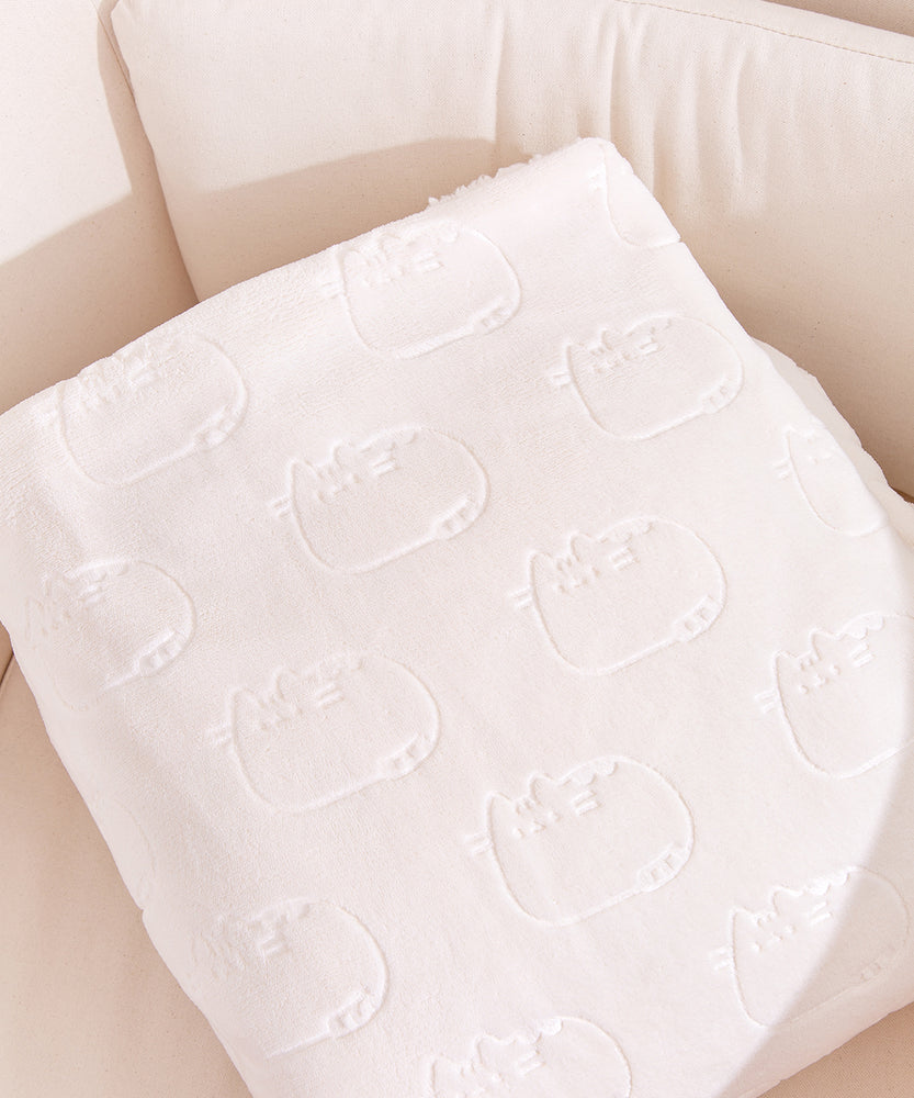 Close-up of the repeating outline print of the Pusheen Blanket. The embossed graphic shows Pusheen lounging with a smile on her face. The cream blanket is folded and set upon a cream-colored chair. 